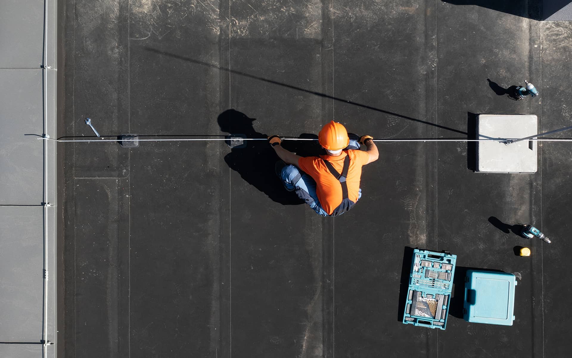 Top-down view of a construction worker using safety equipment while working on a black commercial roof.