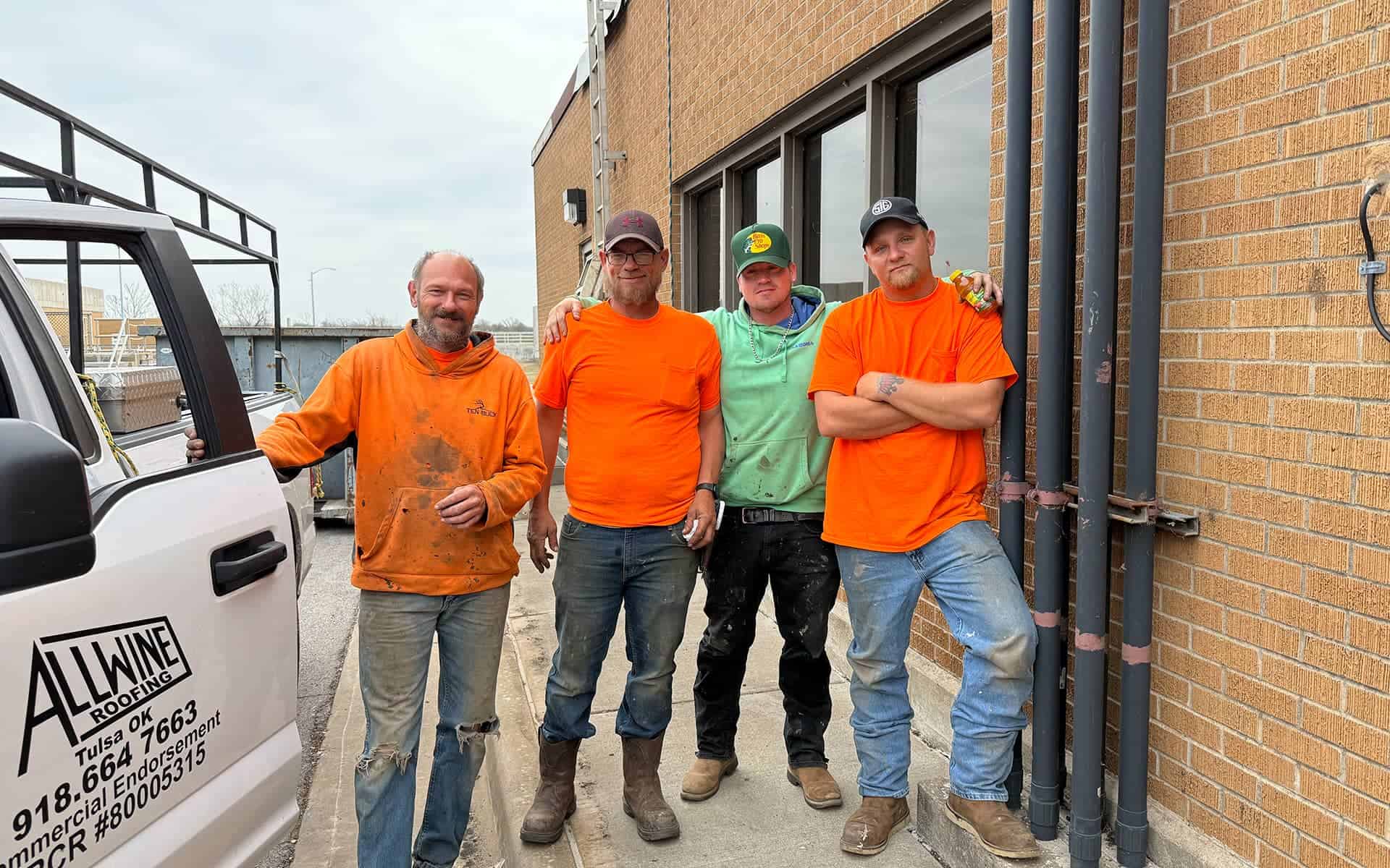 Four Allwine Roofing team members wearing bright shirts, posing together beside a white company truck against a brick building.