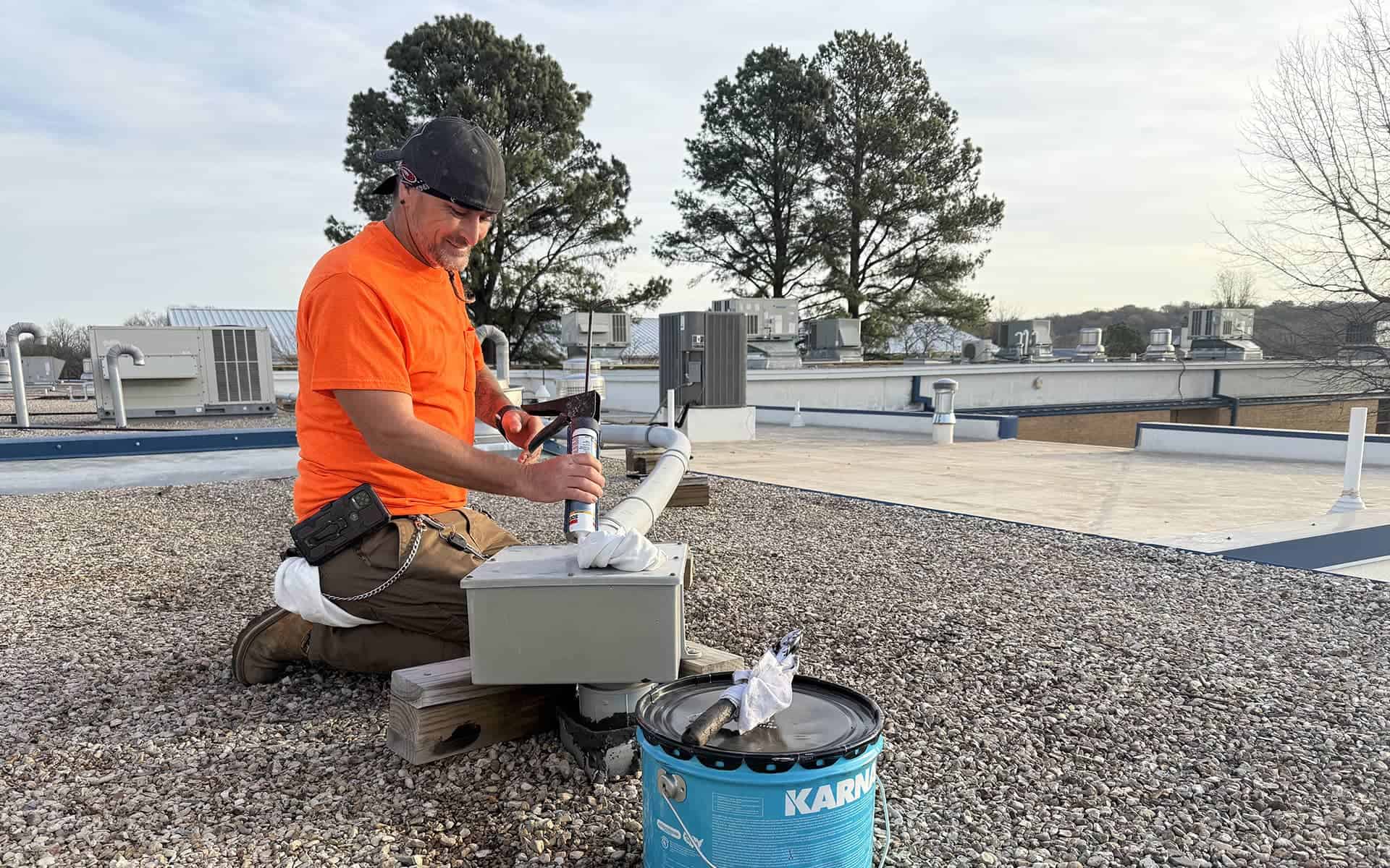 Roofer in an orange shirt using a caulking gun to apply sealant on a rooftop box, with a can of roofing product nearby.