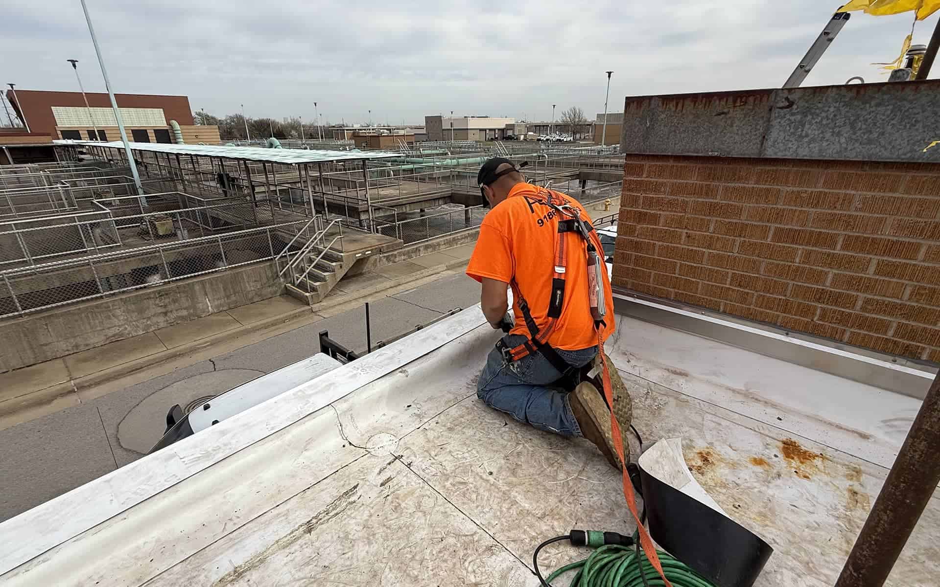Roofer in an orange shirt and harness kneeling at the roof’s edge, fastening or sealing membrane against a brick parapet.