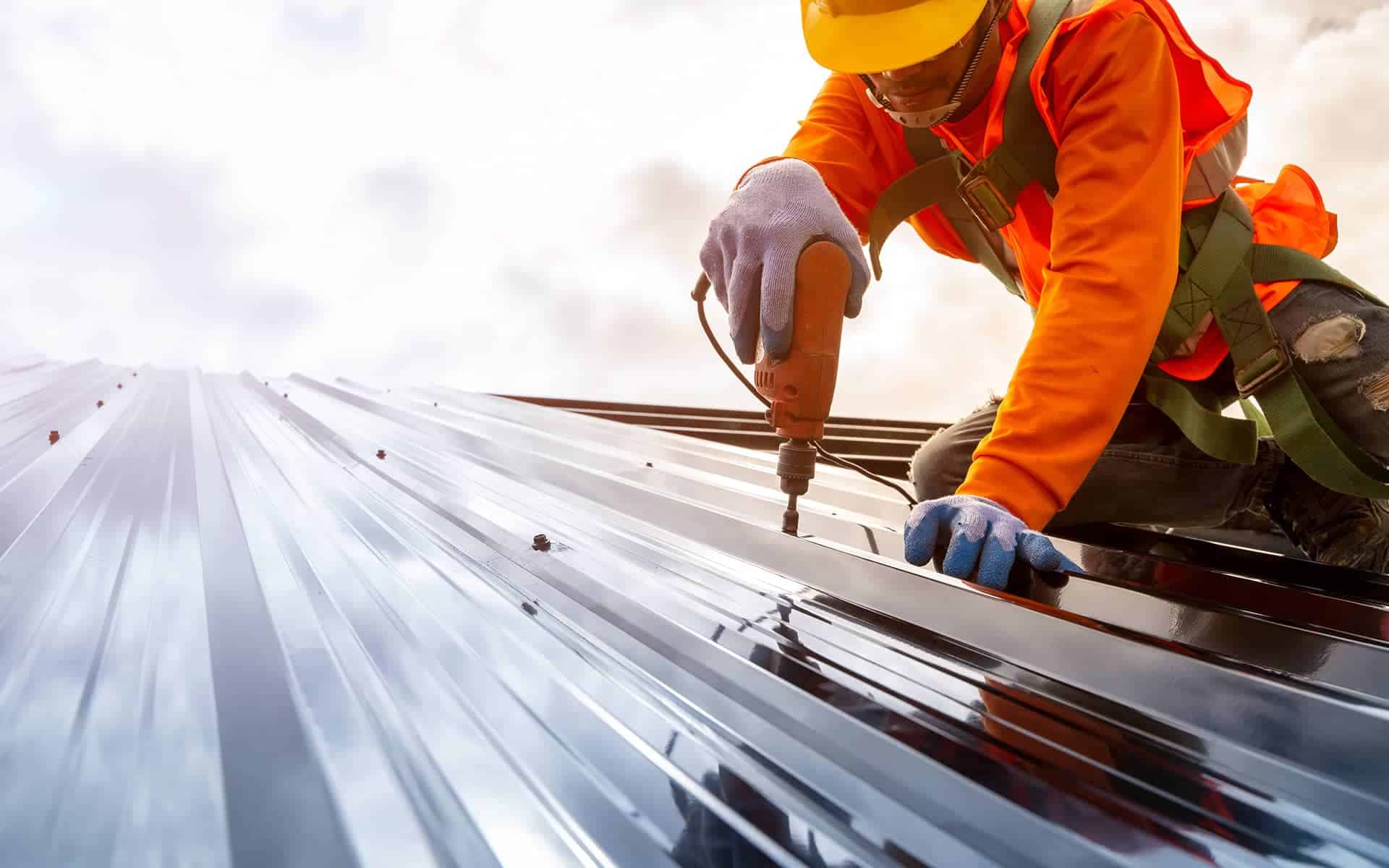 Roofer in protective gear drilling fasteners into a shiny metal roof panel.