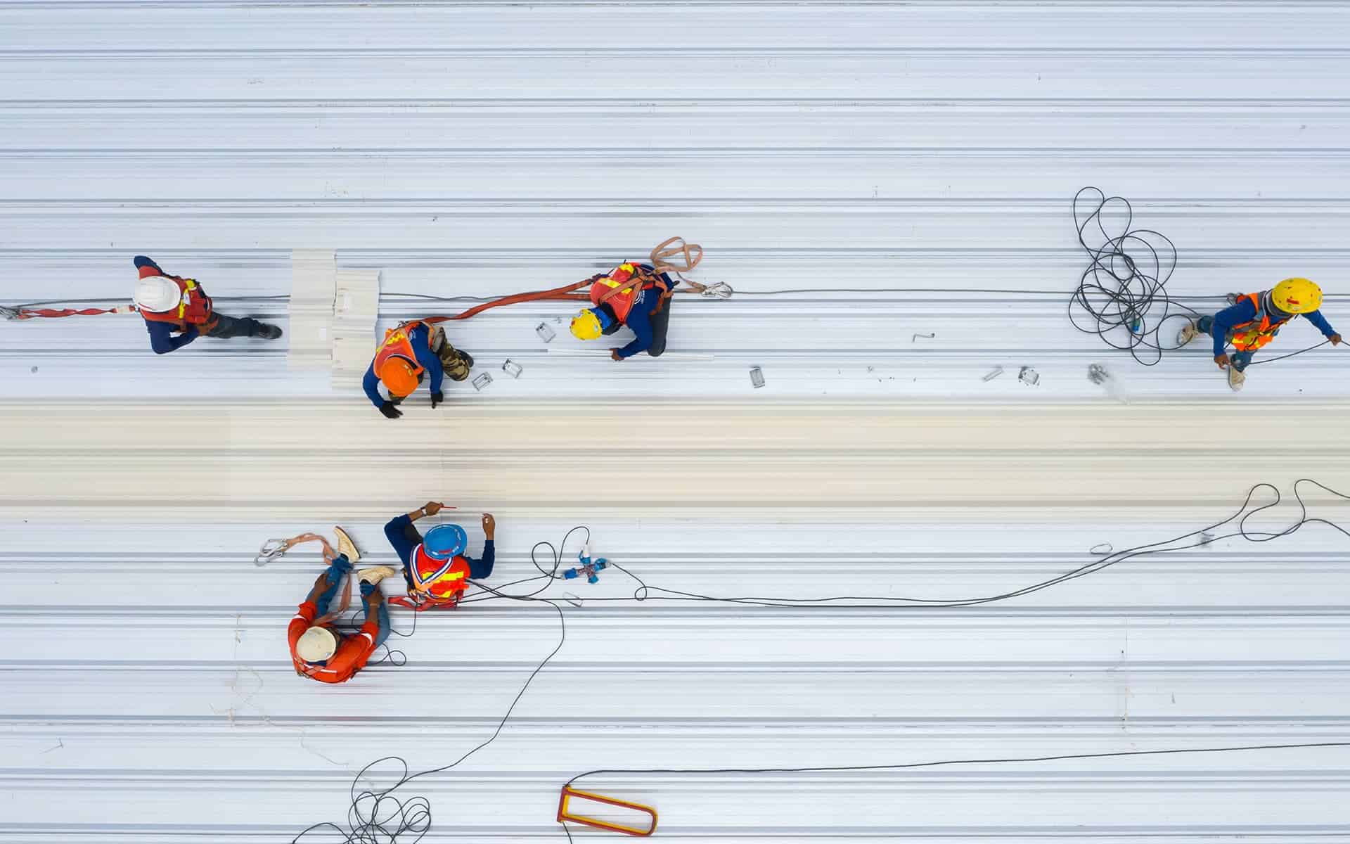 Top-down view of a crew in safety harnesses installing or repairing sections of a white metal roof.